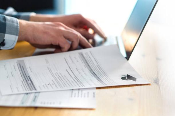 Young man writing college or university application form with laptop. Student applying to school. Scholarship document, admission paper or letter on table. Typing email. Education and communication.